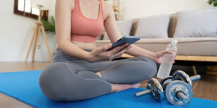 Woman sit on mat holding phone watching online workout training tv class video, using fitness app at home