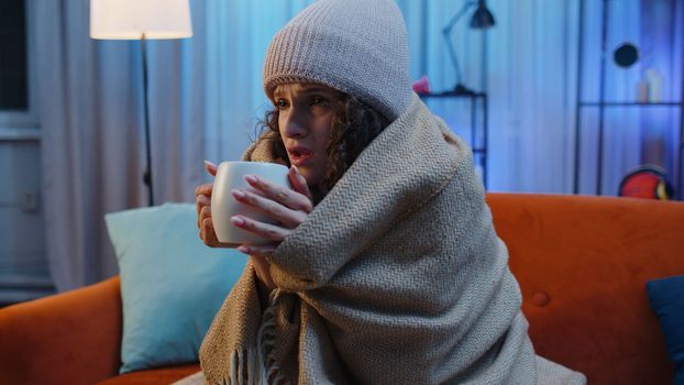 Sick unhealthy woman wear hat wrapped in plaid sit, shivering from cold on sofa drinking hot tea