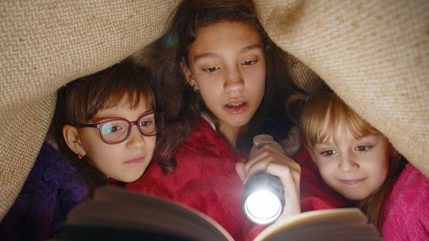 Sisters girl kids under blanket reading interesting fairytale story book using flashlight at home