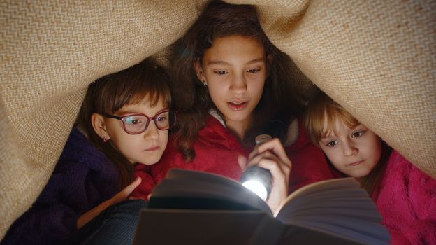 Sisters girl kids under blanket reading interesting fairytale story book using flashlight at home