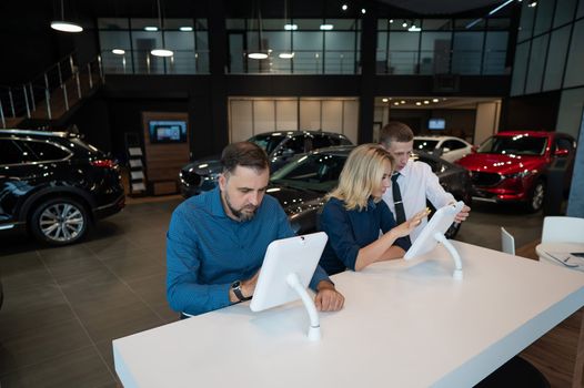 Caucasian married couple chooses a car in a car dealership on digital tablets.