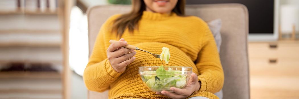 Happy pregnant young woman sitting and eating salad at home