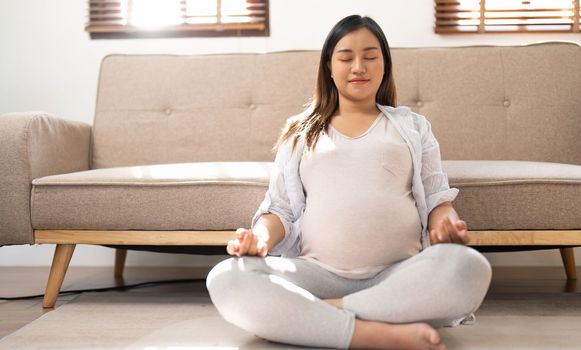 Relaxed Asian pregnant woman meditating in her living room, lotus pose, concentrating breath