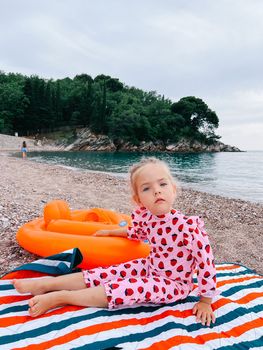 Little girl sits on a blanket on a pebble beach with her hand on an inflatable ring
