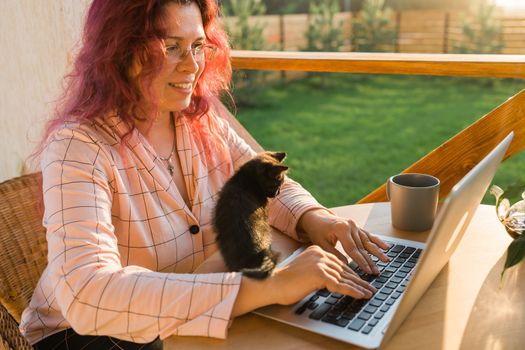 Work from home or study online and videocall with a woman gardener working at laptop outdoor on terrace with kitten, sitting on ratang chair around houseplant and flower pots