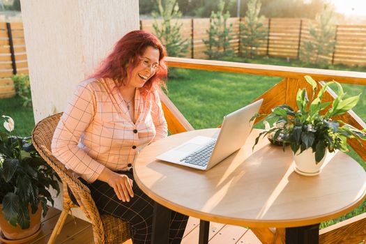 Laughing woman wearing pajama having video call on laptop during lunch break sitting at terrace in her home - summer time and video chat concept