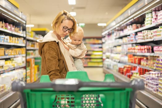 Mother shopping with her infant baby boy child, pushing shopping cart down department aisle in supermarket grocery store.