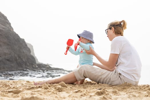Mother playing his infant baby boy son on sandy beach enjoying summer vacationson on Lanzarote island, Spain. Family travel and vacations concept.