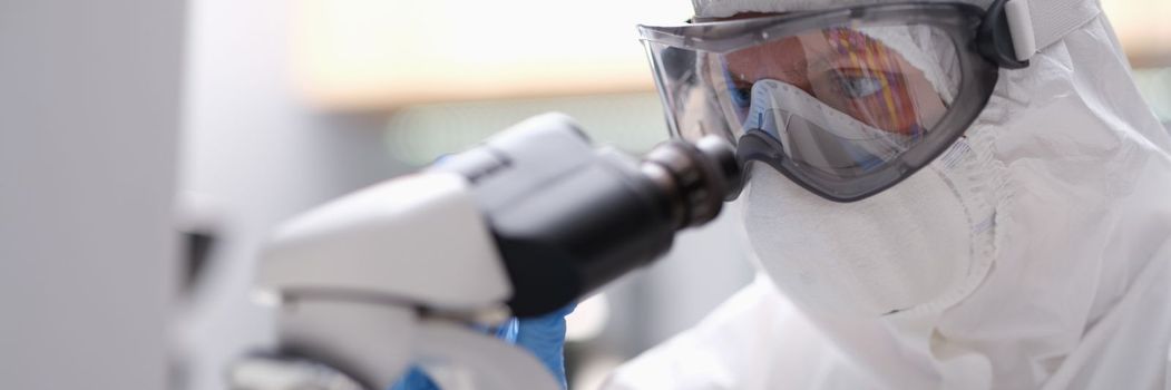 Biologist in protective suit with goggles and medical mask working with microscope in laboratory