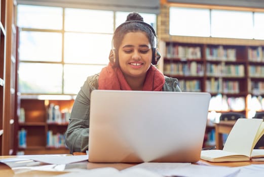 She goes in study mode once she puts her headphones on. a cheerful young female student working on a laptop while listening to music with her headphones inside of a library.