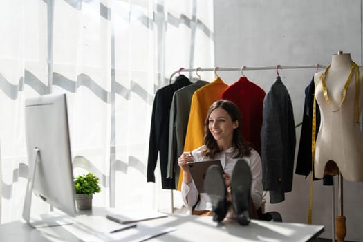 Full length portrait of attractive smiling young stylish fashion clothes designer sittng near table with computer, enjoying working in modern showroom atelier, casual workday concept.