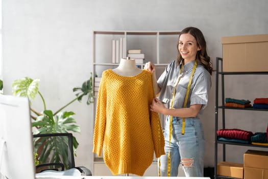 Female clothes designer working with new woman wear collection in cozy workshop studio, fashion designer, dressmaker standing near clothes rack, touching, holding in hands fashionable handmade clothes