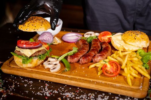 Process of cooking burgers. Cropeed view of chef hands in black gloves with prepared cheeseburger, variety of fillings and ingridients on wooden desk. Catering and Good Quality Fastfood concept