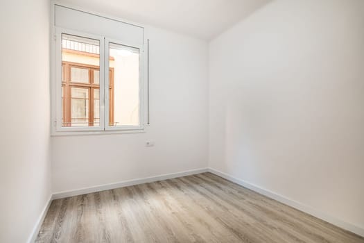 An empty bright room with a window in which the windows of a nearby house are visible. External metal shutters are built into the window to darken the room during daylight hours.
