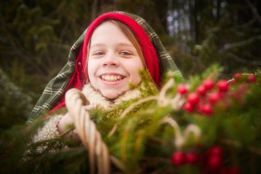Nice cute teenager girl in old peasant clothes with hot fur coat and shawl in cold winter forest with fir trees, pines and snow on winter day. Child with basket full of green branches and red berries