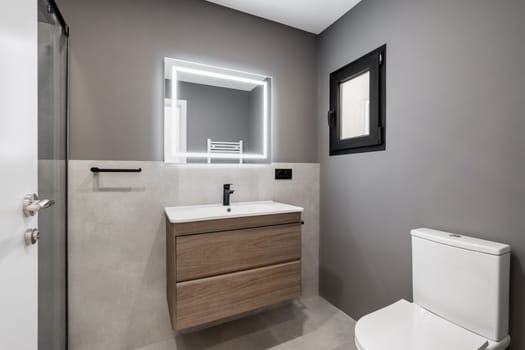 Part of the bathroom in gray and white pastel colors with a matte white ceiling. Sink on a wooden countertop with drawers. On the tiled floor against the wall is a white toilet with a closed lid.