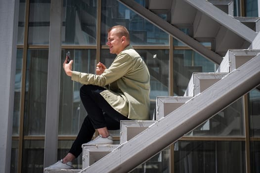 Young man sitting on the stairs and talking sign language via video link on smartphone outdoors.