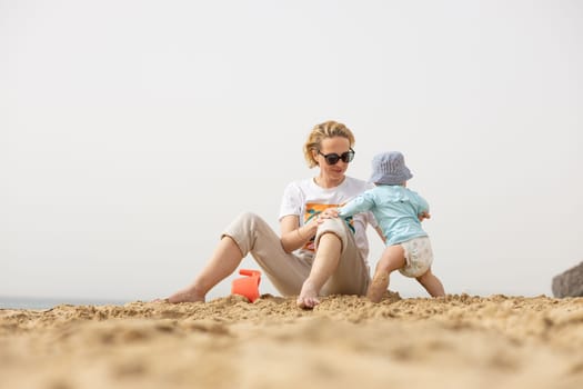 Mother playing his infant baby boy son on sandy beach enjoying summer vacationson on Lanzarote island, Spain. Family travel and vacations concept