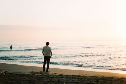 Young man stands on the beach and looks at the sunset. Back view