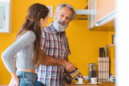 Senior man looking at young woman standing next to him, while he makes coffee for the both of them