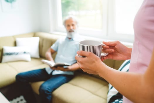 Unrecognizable woman holding a cup of the for her grandfather sitting down on the couch