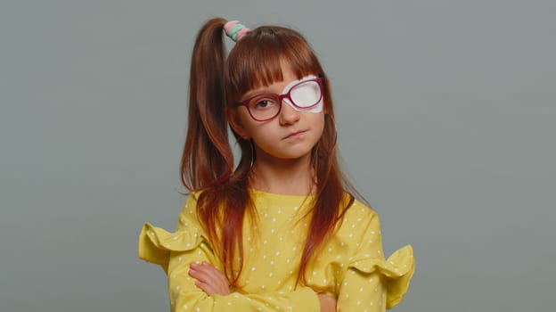 Sick ill preteen girl looking at camera having protective bandage plaster patch scratch on one eye. Injury, trauma, blind. Child kid wearing glasses at gray background. Medical eyesight vision therapy