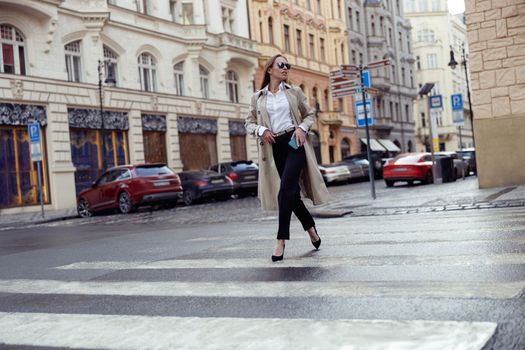 Beautiful woman in trendy outfits crosses road against backdrop of city buildings
