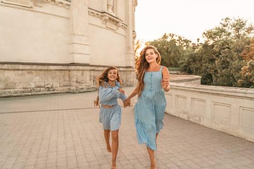 Daughter mother run holding hands. In blue dresses with flowing long hair against the backdrop of a sunset and a white building.