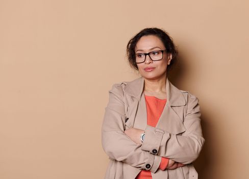 Pensive sophisticated woman wearing eyeglasses, thoughtfully looking aside, posing with folded arms on cream background