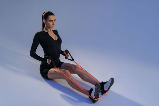Focused woman sit on floor and performs fitness exercises with resistance band on studio background