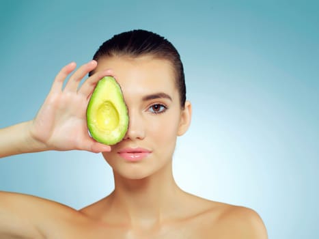 Chemical free and completely natural. Studio portrait of a beautiful young woman covering her eye with an avocado against a blue background.