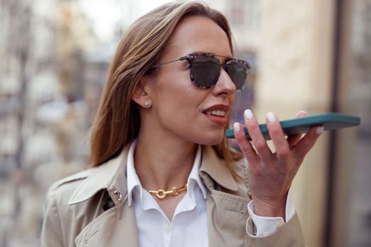Happy businesswoman in sunglasses is recording audio message on phone on city street background