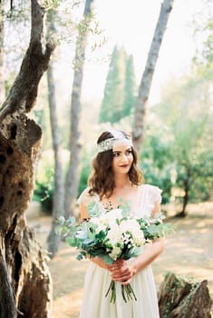Bride with a bouquet in her hands stands under an old olive tree