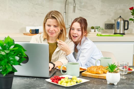 Happy mother and daughter having breakfast in kitchen and using digital devices. Lifestyle