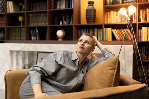 stylish authentic caucasian mature woman with a short haircut sits glorified in an easy chair against the backdrop of shelves with books and vases
