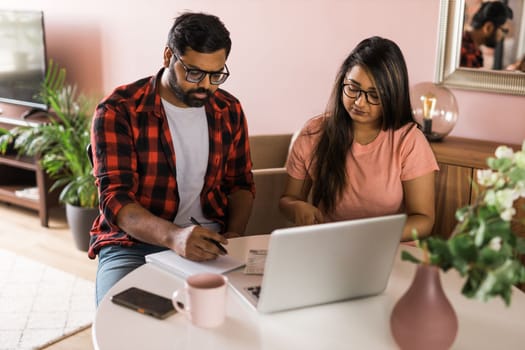 Young millennial indian husband and wife doing domestic paperwork, accounting job and reviewing paper bills, receipts at laptop computer, using online calculator and paying mortgage rent fees on internet - economic crisis concept