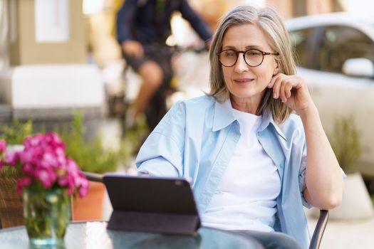 mature European woman seated at an outdoor cafe, browsing information on her tablet PC. She appears relaxed and content, enjoying her leisure time while staying connected through the convenience of technology. The photo captures the essence of modern communication and leisurely lifestyle