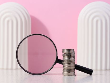 A stack of metal coins and a plastic magnifier on a pink background. The concept of increasing taxes, subsidies