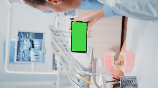 Vertical video: Dentist looking at mobile phone with green screen in dentistry office. Woman working with isolated background and mockup template on smartphone for stomatological care