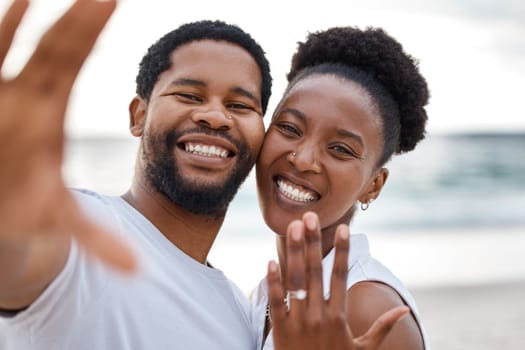 Portrait of an african american couple taking a selfie while showing a ring at the sea together. Happy boyfriend and girlfriend taking a photo after eating engaged on the beach