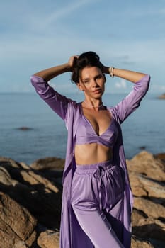 Young beautiful woman in a romantic mood, lilac silk clothes, on the beach against the backdrop of the sea and stones at sunset