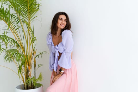 Stylish young woman with voluminous hair in a trendy long sleeve crop top and a pink skirt posing against a white wall and a tropical bush, wearing a smart watch