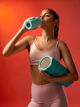 Quenching her thirst after a good session. Studio shot of a sporty young woman drinking water and holding a foam roller against a red background.