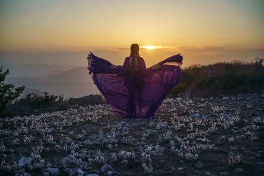 Sunset purple dress woman mountains. Rise of the mystic. sunset over the clouds with a girl in a long purple dress. In the meadow there is a grass dream with purple flowers.