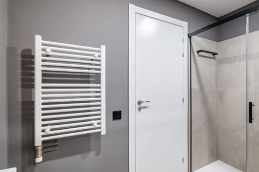 Part of a bathroom with a gray wall. White front door with metal handle. On the wall is a white radiator for drying towels. The shower area is enclosed by sliding glass doors.