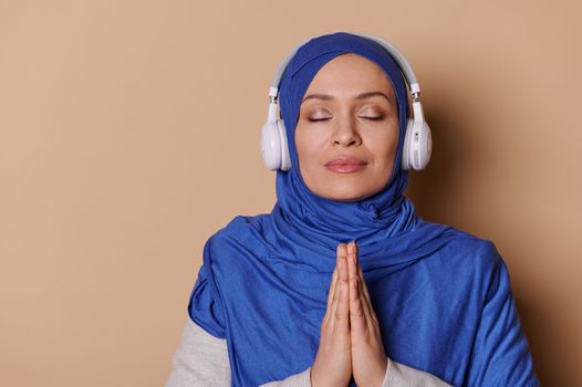 Arab Muslim woman with her eyes closed, in a hijab, keeps hands palms together, listens to soothing music on headphones