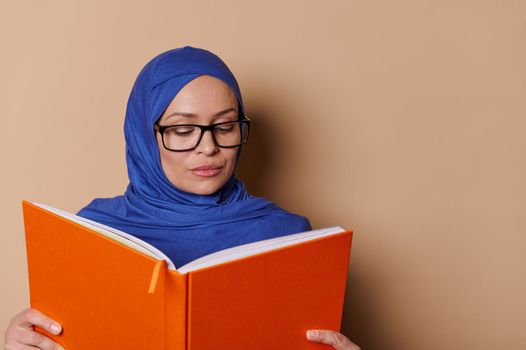 Educated sophisticated Muslim woman smiling at camera while reading book, standing isolated on a beige cream background