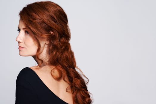 Cascading curls down her back. Studio shot of a young woman with beautiful red hair posing against a gray background.