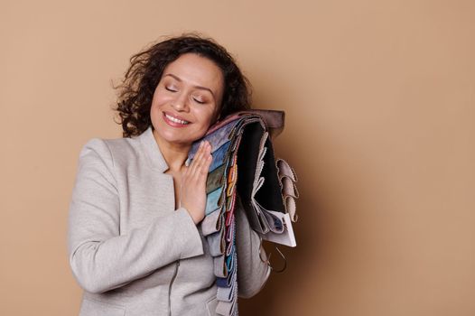 Charming woman testing fabric samples for softness, smiling a toothy smile, posing with eyes closed on beige background