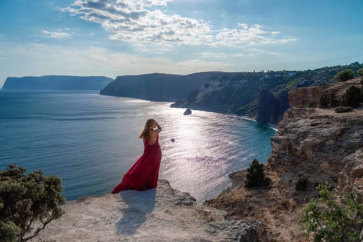 A girl with flowing hair in a long red dress stands on a rock above the sea. The stone can be seen in the sea. Sunny path to the sea from the sun.